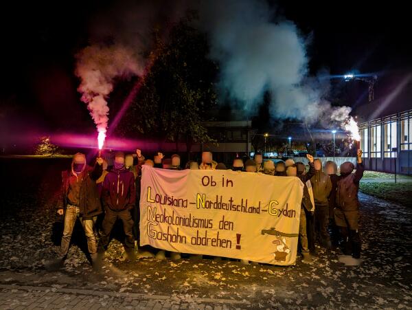 Das Bild wurde im dunkeln auf einer Wiese aufgenommen. Ca 20 Menschen stehen im Vordergrund. Die Menschen halten ein Transpi hoch auf dem steht Ob in Luisiana, Norddeutschland, Gydan, Neokolonialismus den Gashahn abdrehen. Auf dem Transpi ist in der rechten unteren Ecke ein gemalter Hase der einen Gashahn abdreht. 