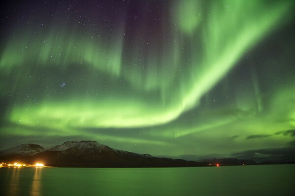 A night sky absolutely filled with green, glowing, dancing aurora. They reflect in a now green ocean and cast their glow against snow top mountains. 