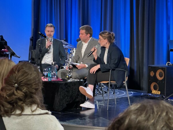 Eine Podiumsdiskussion mit drei Rednern, die auf der Bühne sitzen, wobei einer ein Mikrofon in der Hand hält, während die anderen sich unterhalten. Die Einstellung enthält Musikgeräte im Hintergrund und ist mit blauem Licht beleuchtet. Die Zuschauer sind im Vordergrund teilweise sichtbar.