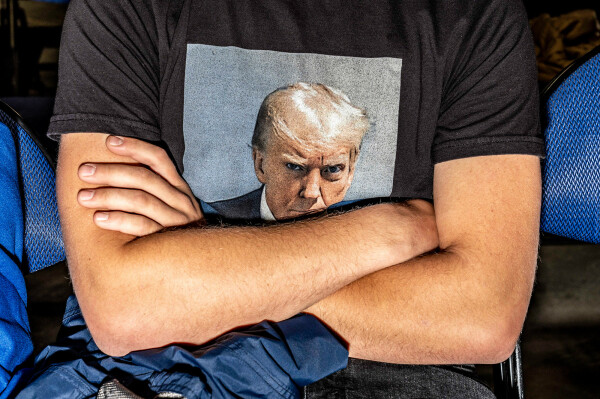 Zu sehen ist der Oberkörper einer Person mit verschränkten Armen, die ein Trump-T-Shirt trägt.