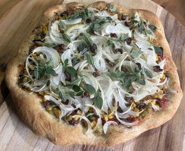 A freshly baked pizza topped with thinly sliced onions, fresh organo, peas, corn and black olives, resting on a wooden surface.