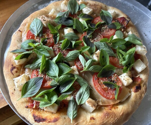 A freshly baked pizza topped with fresh basil leaves, sliced tomatoes, and vegan cheese cubes, on a round metal pizza tray.