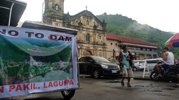 zu sehen ist ein Gebäude mit Bergen im Hintergrund , davor ein Auto vor welchem ein Mensch läuft. Daneben ein Banner (ist nur halb zu sehen)

Auf dem Banner zu sehen ist nur ein Teil der BEschriftung " O Dam" darunter Bild einer Lagune "Lagura" steht darunter